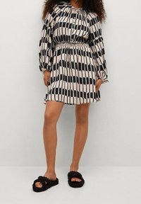 Vestido impreso con rayas verticales en negro y crema, mangas largas abullonadas, cintura fruncida y cuello. Combinado con sandalias deslizantes trenzadas en negro.
