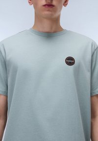 T-shirt azzurro chiaro a maniche corte realizzato in tessuto morbido, con una toppa circolare nera bordata di arancione sul petto.