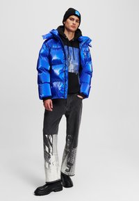 Giacca a vento blu lucido con imbottitura in piuma, felpa nera sotto, jeans grigi con stampa grafica e stivali neri chunky.
