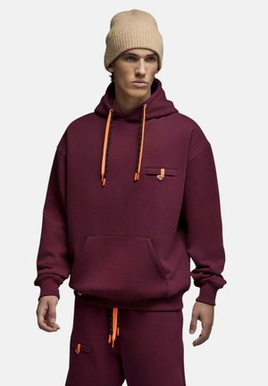 Jeune homme portant un bonnet en tricot beige et un sweat à capuche bordeaux avec des cordons orange, assorti à un pantalon de survêtement bordeaux.