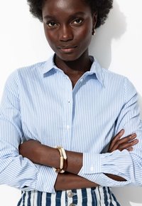Chemise à rayures bleues et blanches, à boutons, avec un col, des manches longues et des poignets ; portée avec des bracelets dorés et bruns sur des bras croisés.