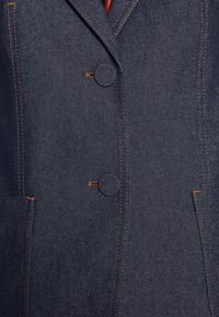 Veste en denim foncé avec des coutures orange visibles, deux boutons recouverts de tissu et deux poches verticales à l'avant, présentée de la taille à la tête.