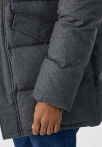 Giacca imbottita grigia con tessuto strutturato, caratterizzata da una manica sagomata e tasche laterali, indossata con pantaloni di denim blu e una mano visibile.