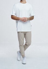 Muž s tetovanými pažemi, oblečený v bílém oversize tričku, béžových úzkých kalhotách a bílých teniskách, stojící se zkříženýma nohama.
