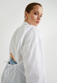 Hvid skjortejakke med lange ærmer, der har en samlet ryg og justerbare snore. Afsluttet med en glat tekstur og et afslappet design.