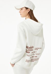 Weiße Kapuzenjacke mit Kapuze, die auf der Vorderseite in dunkelbrauner Schrift "Manchester United Kingdom 1998" und Veranstaltungsinformationen auf der Rückseite aufweist. Weiße Mütze.
