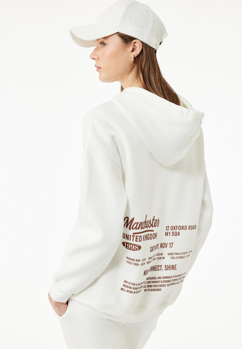 Weiße Kapuzenjacke mit Kapuze, die auf der Vorderseite in dunkelbrauner Schrift "Manchester United Kingdom 1998" und Veranstaltungsinformationen auf der Rückseite aufweist. Weiße Mütze.