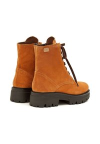 Botas de tobillo de ante marrón con una punta redonda, suelas gruesas negras y cordones marrones. Incluye una pequeña etiqueta de marca en el lateral.