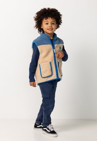 Kleines Kind mit lockigem Haar, das eine blaue und beige Fleece-Weste über einem dunkelblauen Shirt und einer Hose trägt, mit schwarzen Sneakers, stehend vor einem weißen Hintergrund.