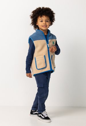 Jong kind met krullend haar, gekleed in een blauw en beige fleece vest over een marineblauw shirt en broek met zwarte sneakers, staand op een witte achtergrond.