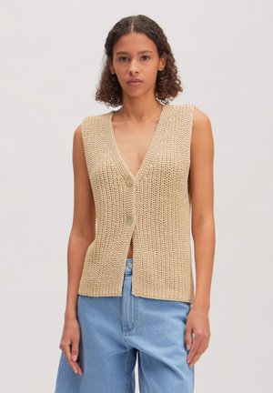 Vrouw met krullend haar, die een mouwloos beige gebreid vest met twee knopen draagt, gecombineerd met lichtblauwe high-waisted jeans, staand tegen een effen achtergrond.