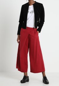 Svart mockajacka med dragkedja fram och figurnära design, vit grafisk t-shirt under, röda vida byxor och svarta snörskor.