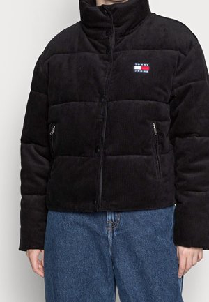 Person trägt eine schwarze Cord-Steppjacke mit Tommy Jeans-Logo und blaue Jeans, steht vor einem einfarbigen Hintergrund.