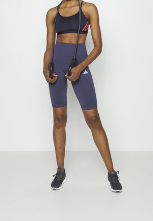 Laivastonsiniset korkeavyötäröiset pyöräilyshortsit, joissa on pehmeä pinta ja istuva muotoilu, yhdistettynä mustaan urheiluliiviin, jossa on punaiset yksityiskohdat, ja hyppynaru.