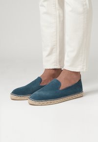 Des chaussures slip-on en daim teal avec une semelle ext�érieure en corde de jute, présentant un bout rond et un dessus sans embellissement. Portées avec un pantalon blanc à jambes courtes.