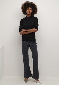 Suéter negro de manga corta combinado con jeans acampanados oscuros. La modelo se mantiene erguida y segura, vistiendo tacones plateados de punta afilada. Fondo minimalista.