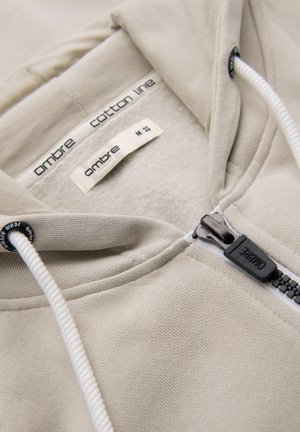 Beżowa bluza z kapturem na suwak z białymi sznurkami, ocieplana polarem, z metką „ombre cotton line” wewnątrz kołnierza, rozmiar średni.