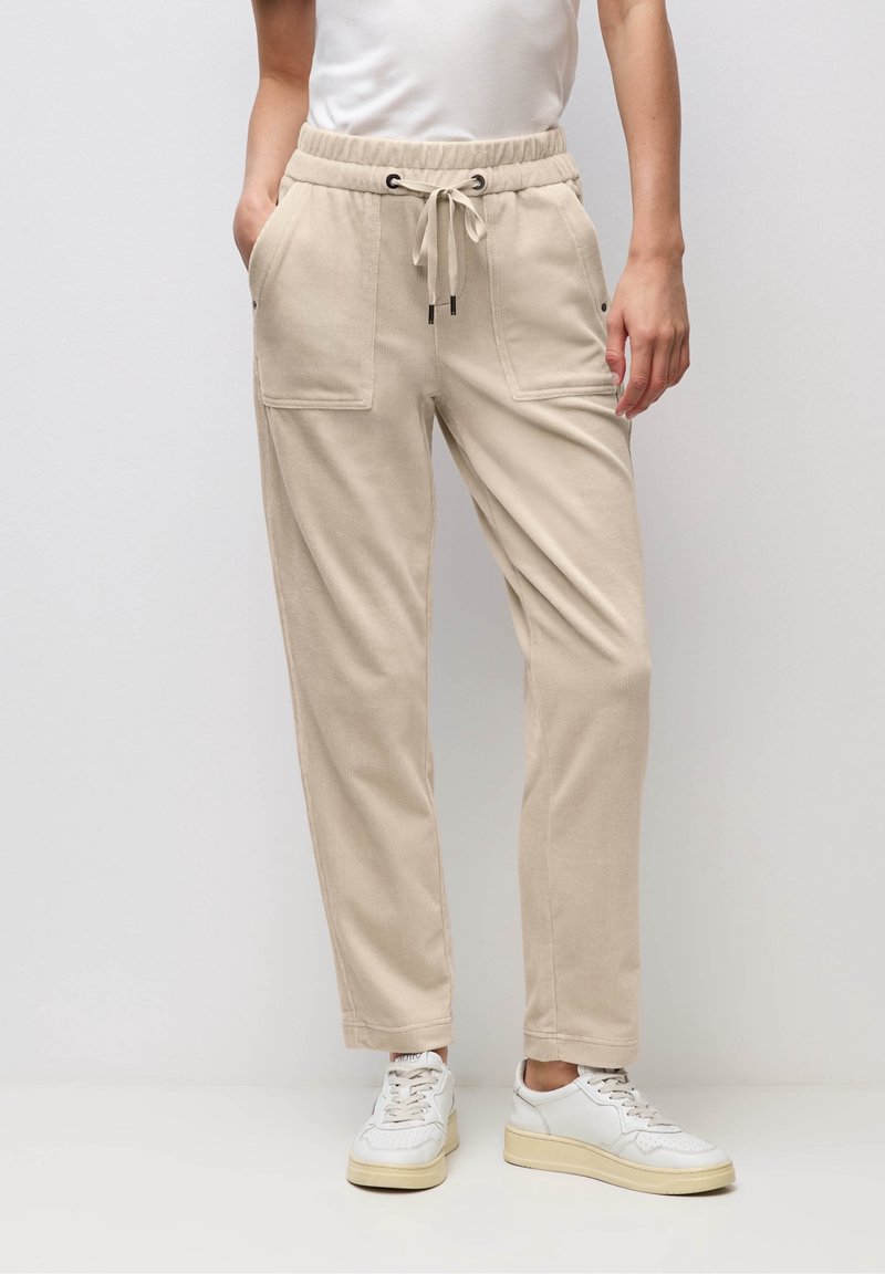 Personne portant un pantalon beige à cordon avec poches plaquées à l'avant et des baskets blanches, debout devant un fond clair uni.