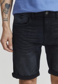 Mørke denimshorts med optrukne bukseben, der har en frontlomme og metalknapdetalje. Stoffet har en glat tekstur og subtil falming.
