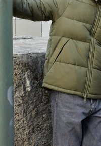 Oliwkowa pikowana kurtka puchowa z zamkiem błyskawicznym, zestawiona z szarymi dżinsami. Tekstura wydaje się miękka i wyściełana.