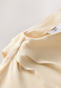 Vêtement sans manches de couleur crème avec un détail de tissu froncé près de l'encolure, montrant une étiquette blanche « NEXT » à l'intérieur.