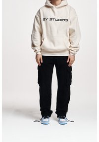 Krémová mikina s kapucí a černým nápisem "2Y STUDIOS", přední kapsa, nošena s černými cargo kalhotami a modrými teniskami se šedými akcenty.