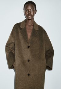 Cappotto di lana marrone con colletto a revene, vestibilità oversize e tre bottoni neri. Texture liscia, senza motivi o decorazioni visibili.