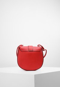 Červená kožená křížová taška s kulatým dnem a klopou, vystavená na bílém podstavci na jednoduchém světlém pozadí.