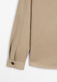 Camicia beige a maniche lunghe con colletto classico, polsini abbottonati e una superficie liscia. Presenta una vestibilità su misura e dettagli sulle cuciture laterali.