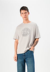 Camiseta de gran tamaño en gris claro con escote redondo y un gráfico central impreso que dice "GARMENTOS DE CALIDAD SUPERIOR". Los jeans de mezclilla tienen un corte recto.