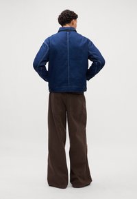 Donkerblauwe denimjacket met een kraag en contraststiksels, gecombineerd met losse, bruine wijde broeken met achterzakken.