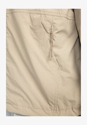 Chaqueta ligera beige con un diseño de panel horizontal, costuras visibles y un bolsillo con cremallera y tirador en el lateral.