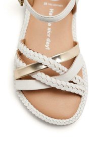 Sandal blanc avec des brides tressées et un accent doré. La semelle intérieure présente une couleur beige clair avec le texte : "Bonne journée !"