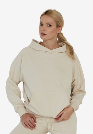 Kremowy hoodie wykonany z miękkiego materiału, z dużą kieszenią z przodu i kapturem ze sznurkiem. Luźny krój z ściągaczami u rękawów.