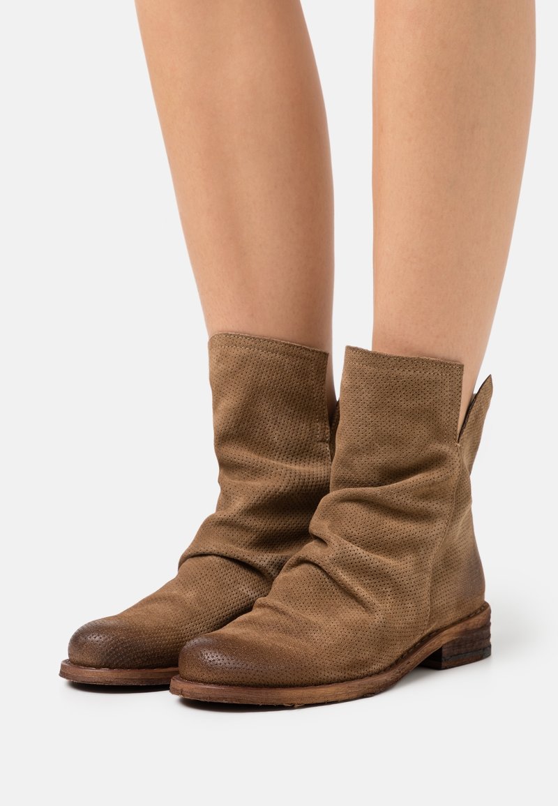 Bottines en daim marron clair avec un design ample et des petits talons en bois portées sur des jambes nues sur un fond blanc.