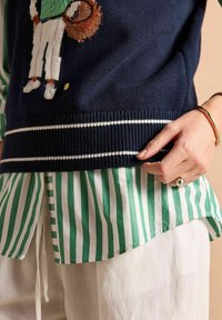 Tumes sinine kootud sviiter, millel on tikitud tennisemängija, millele lisab valgetriibuline soonik. All on roheline ja valge triibuline särgis.