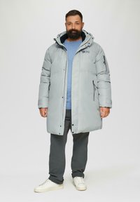 Lichtgrijze, gewatteerde jas met een capuchon, die zwarte ritszakken en accenten heeft. Draag het over een blauwe trui en grijze broek, gecombineerd met witte sneakers.