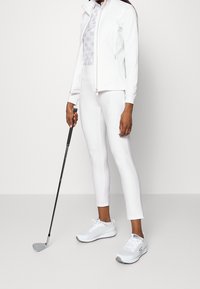 Kobieta w białym stroju sportowym trzymająca kij golfowy, stojąca na tle jasnym.