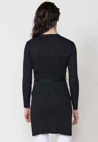 Vestido negro de punto acanalado con mangas largas, una silueta ajustada y una cintura ceñida. Presenta un escote redondo y sutiles aberturas laterales en el dobladillo.