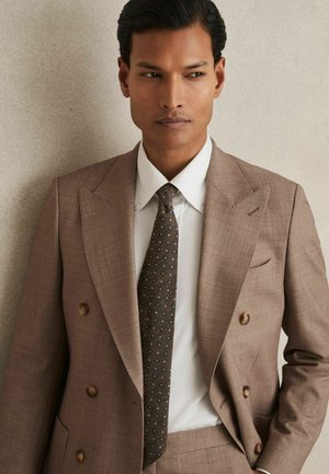 Costume marron à double boutonnage avec un tissu texturé, chemise blanche et cravate sombre à motifs avec des accents bleus.