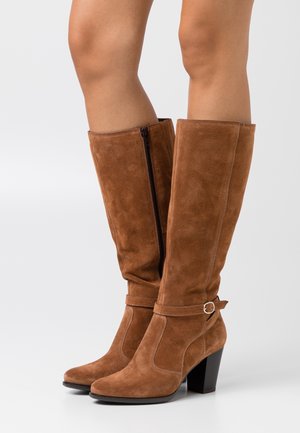 Bottes hautes jusqu'au genou en daim marron avec talons blocs, fermeture éclair côté intérieur et sangle décorative avec boucle autour de la cheville, portées sur des jambes nues.