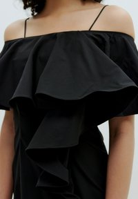 Schwarzes schulterfreies Kleid mit großem Rüschenausschnitt, schmalen Trägern und glatter Stoffoberfläche. Nahaufnahme des Oberteils.