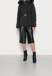 Doudoune noire avec capuche en fourrure, taille ceinturée et fermeture éclair ; associée à un pantalon large en simili cuir noir et des bottines à talons.