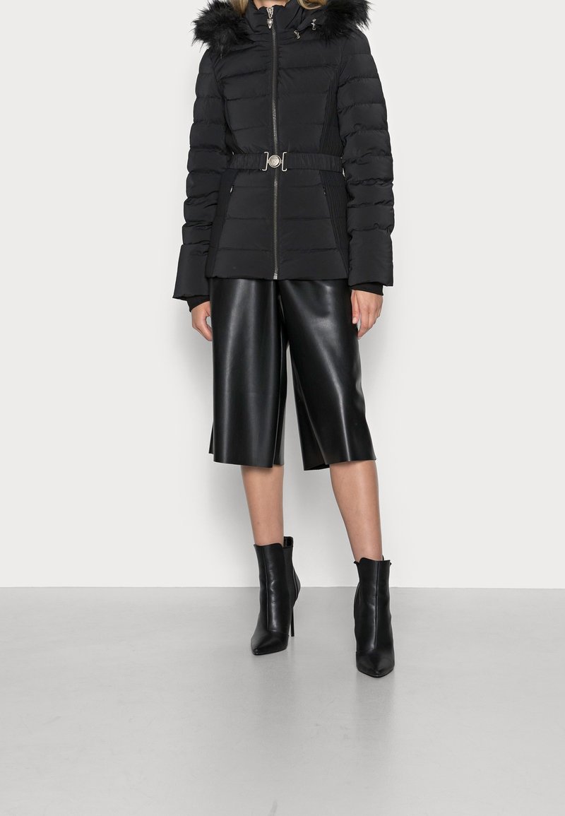 Doudoune noire avec capuche en fourrure, taille ceinturée et fermeture éclair ; associée à un pantalon large en simili cuir noir et des bottines à talons.