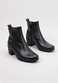 Bottines en cuir noir avec un bout pointu, un talon carré et une fermeture éclair latérale. Présente une sangle décorative et un détail de boucle. Texture lisse.