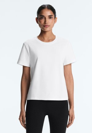 Žena s tmavými vlasmi zviazanými dozadu, oblečená v jednofarebnom bielom tričku s krátkym rukávom a čiernych nohaviciach, stojaca na svetlom pozadí.