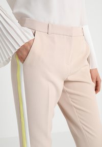 Beige kalhoty na míru s hladkou texturou, zdobené bílými a neonově žlutými bočními pruhy. Kombinované s bílou plisovanou blůzkou s dlouhým rukávem.