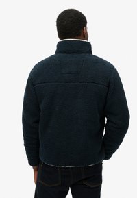 Pile blu navy con collo alto, superficie strutturata e vestibilità comoda. Presenta una cucitura nella parte superiore della schiena.