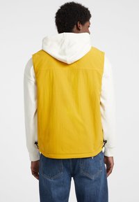 Gelbe ärmellose Weste mit strukturierter Oberfläche, kombiniert mit einem weißen Kapuzenpullover, von hinten gesehen, die einen lässigen Lagenlook zeigt.