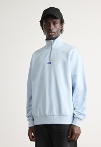 Sudadera de cremallera color azul claro con cuello alto y puños acanalados, que presenta un pequeño logotipo azul en el pecho y un diseño holgado.